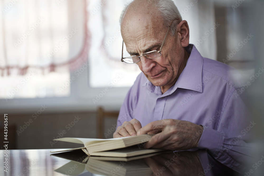 Hoary pensioner wearing glasses sitting at table and reading book Hoary pensioner wearing glasses sitting at table and reading book