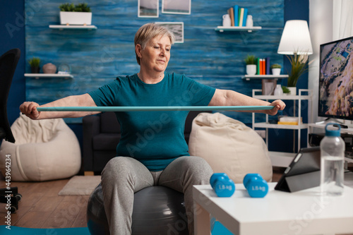 Retired cheerful pensioner sitting on swiss ball watching fitness video on tablet streching arm using elastic band during healthcare workout in living room. Senior woman working body resistance