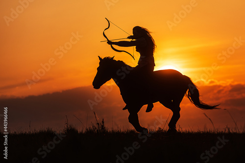 Silhouette against the setting sun woman riding in historical costume as a warrior with bow