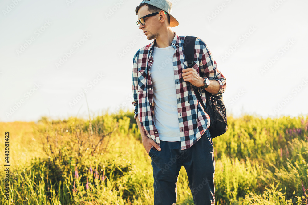 Smiling young man wearing glasses and cap traveling with backpack in summer, hiker hiking and observing picturesque landscape
