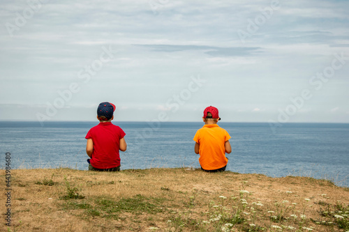 A couple of boys sit on a cliff of the coast and look into the distance into the blue sky.