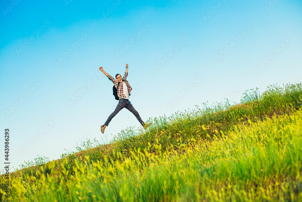 Happy young tourist with a backpack, happy jumping man with hands up against a blue sky background, climbs to the top of a hill or mountain Happy young tourist with a backpack, happy jumping man with hands up against a blue sky background, climbs to the top of a hill or mountain
