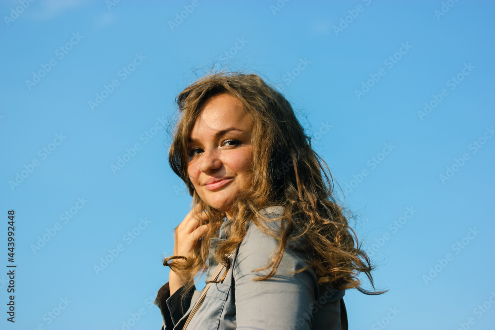 A beautiful swarthy woman with brown curly hair waving in a wind on a sunny day against a blue sky background. Portrait of a happy smiling girl in a gray business suit. A successful businesswoman. A beautiful swarthy woman with brown curly hair waving in a wind on a sunny day against a blue sky background. Portrait of a happy smiling girl in a gray business suit. A successful businesswoman.