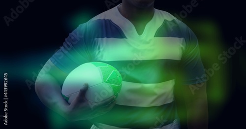 Composition of glowing green blur over male rugby player holding rugby ball, on black