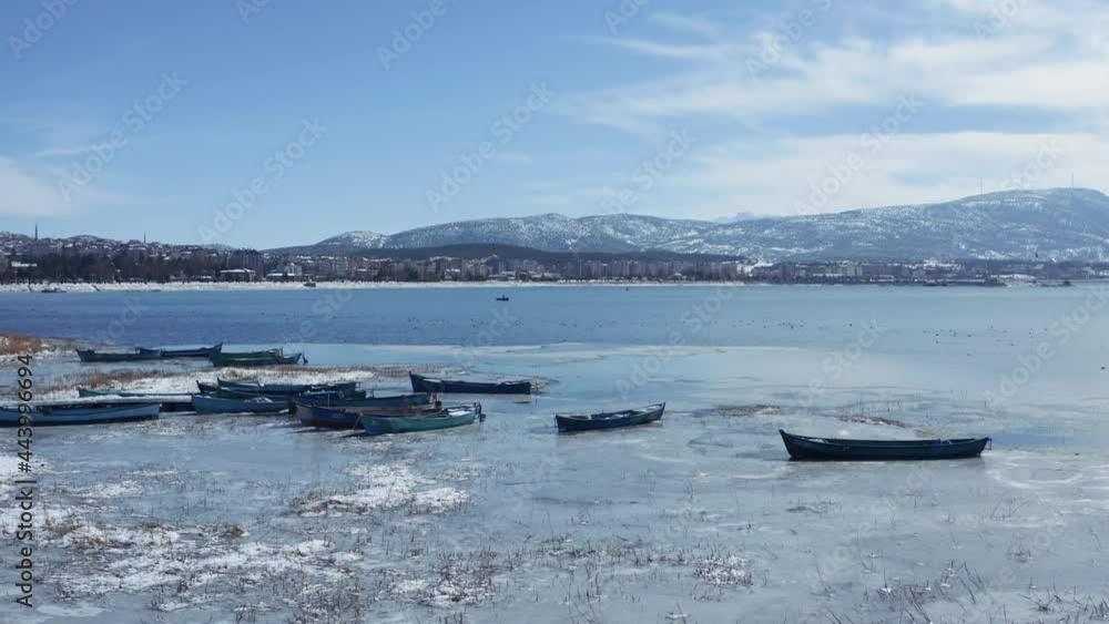 Aerial shot of the half frozen lake and boats docked on the shore on sunny day with city and mountains the in the background. 4K. Aerial shot of the half frozen lake and boats docked on the shore on sunny day with city and mountains the in the background. 4K.