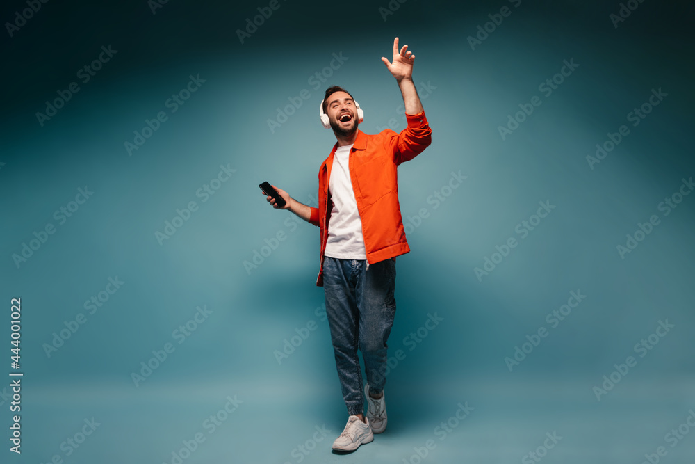 Handsome man wearing jeans and orange jacket listening to music in massive headphones
