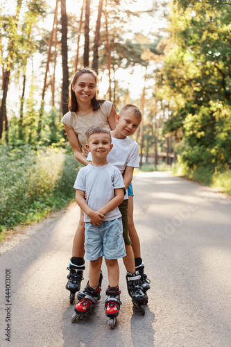 Happy family, dark haired female wearing casual attire standing with her sons outdoor, mother with children roller skating in park on asphalt road, having fun together.