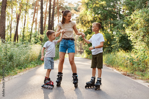 Outdoor shot of happy family having fun and roller skating together in summer park, mommy holding kids hands, being glad to spend weekend together, active pastime.