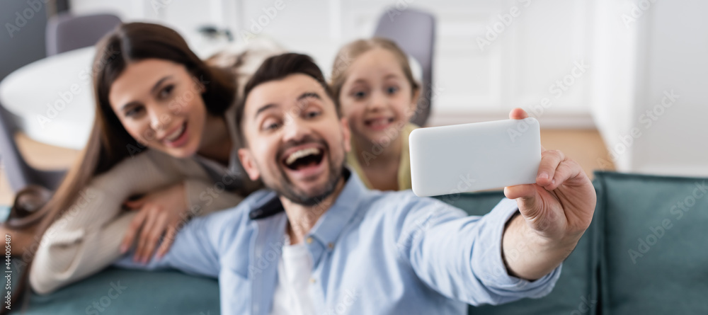 selective focus of smartphone in hand of cheerful man taking selfie with happy wife and daughter, banner selective focus of smartphone in hand of cheerful man taking selfie with happy wife and daughter, banner