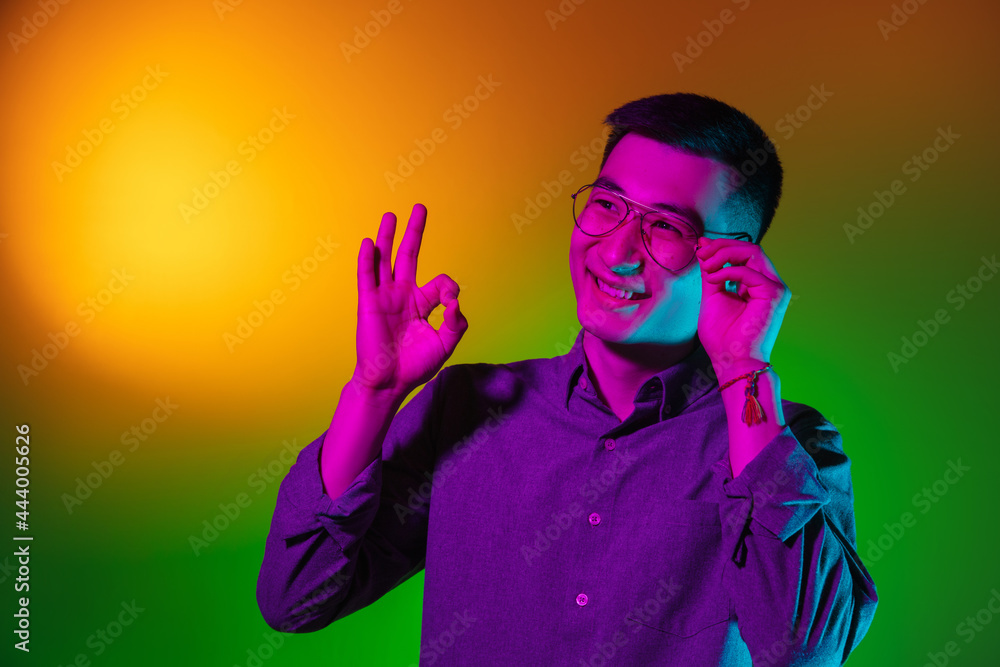 Portrait of Asian young man isolated on studio background in gradient green yellow neon light, colour filter. Concept of human emotions, facial expression.