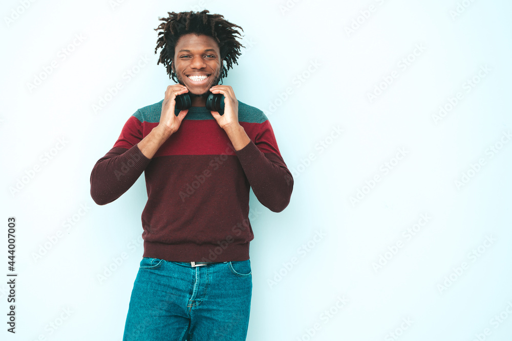 Obraz premium Handsome smiling hipster lambersexual model.Unshaven African man dressed in stylish clothes.Fashion male with dreadlocks hairstyle posing near wall in studio.Listening music in wireless headphones