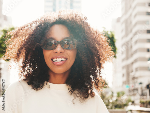Beautiful black woman with afro curls hairstyle.Smiling hipster model in white t-shirt. Sexy carefree female posing on the street background. Cheerful and happy in sunglasses
