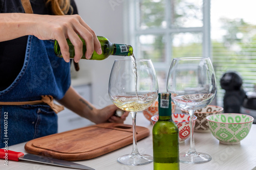 woman in the kitchen pouring two glasses of white wine while she cooks