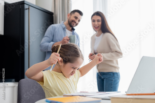 blurred couple smiling near angry girl showing clenched fists while doing homework