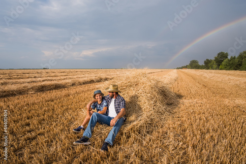 Father and son are resting after successful harvest. Rainbow in the sky behind them.