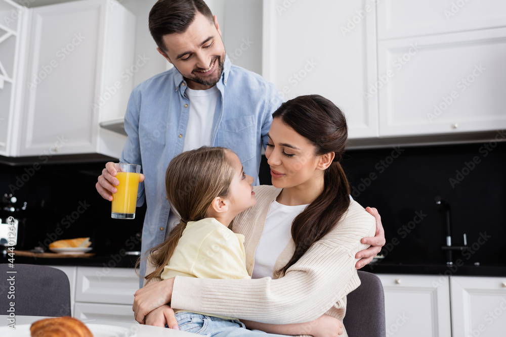 happy woman embracing daughter near husband holding orange juice in kitchen happy woman embracing daughter near husband holding orange juice in kitchen