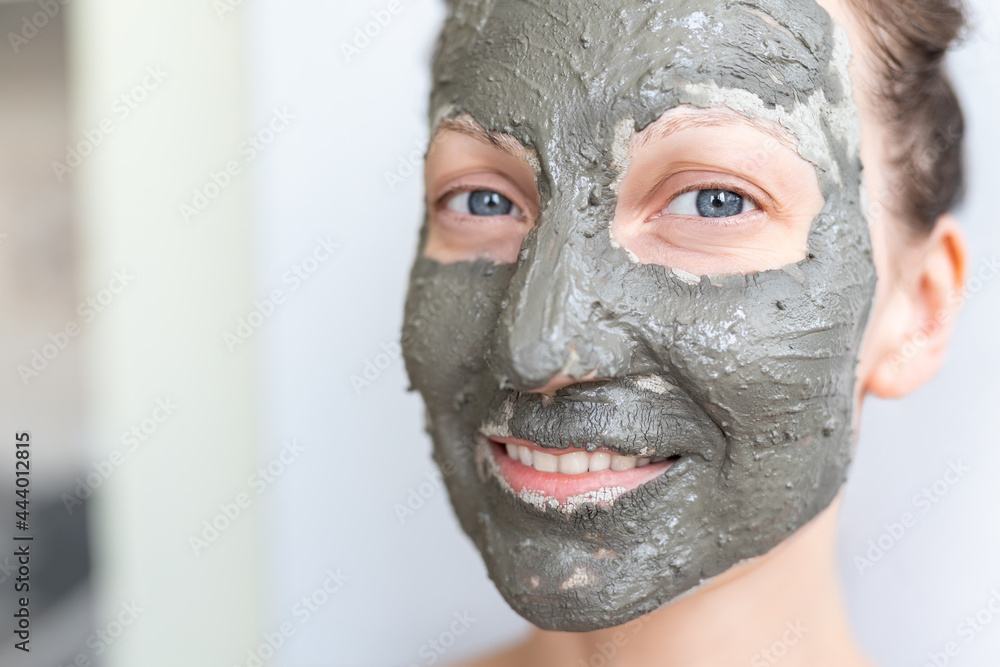 Fototapeta premium Young adult happy beautiful caucasian woman applying natural gray clay face mask at home against window and mirror at day time. Female person making self facial cleansing moisturizing treatment care