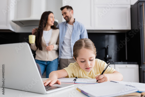 child using laptop and writing in notebook near smiling parents talking on blurred background