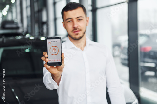 Holds phone with labels and icons. Young man in white shirt is indoors with modern new automobile. Remote control