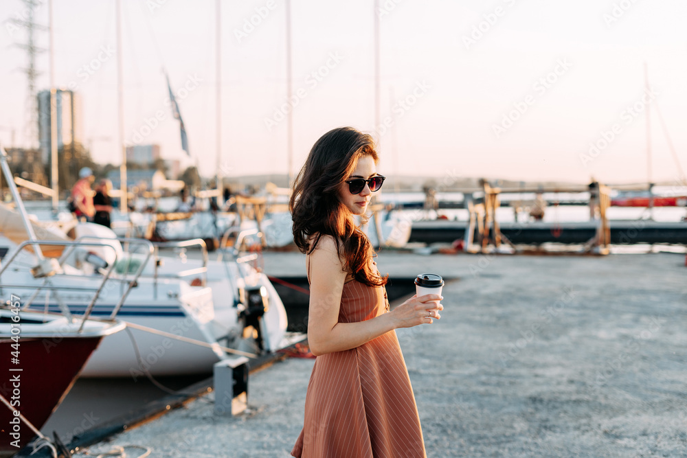 Beautiful young girl walking on the pier with coffee in black glasses in a dress. Sea holidays, life Beautiful young girl walking on the pier with coffee in black glasses in a dress. Sea holidays, life