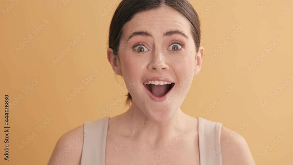 A surprised emotional woman is looking to the camera standing isolated over beige wall in the studio A surprised emotional woman is looking to the camera standing isolated over beige wall in the studio