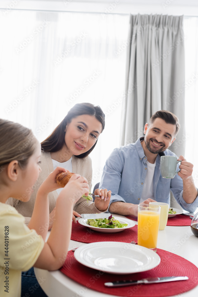 girl eating croissant near happy parents during breakfast girl eating croissant near happy parents during breakfast