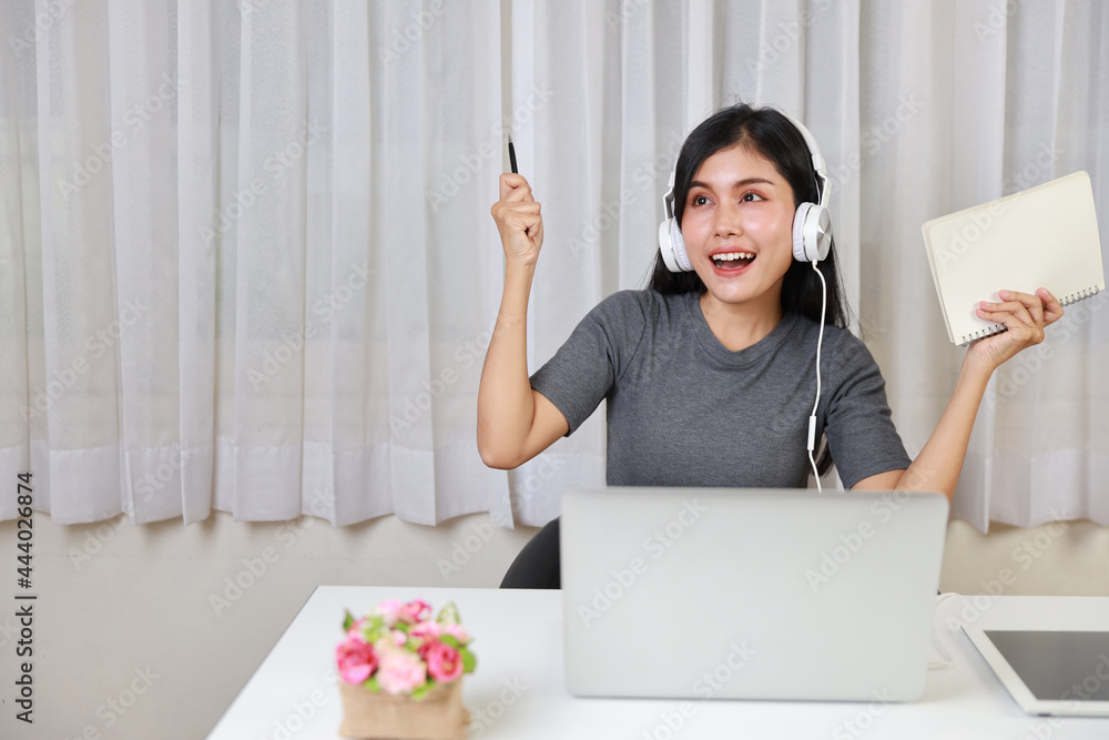 Young smart and active asian female student sitting at table with headset, using computer and taking note for studying online with exciting emotion (new normal concept) Young smart and active asian female student sitting at table with headset, using computer and taking note for studying online with exciting emotion (new normal concept)