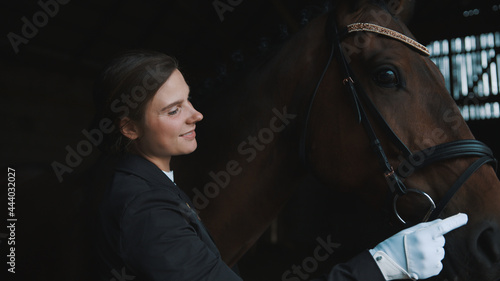 Horsewoman touching the face of her seal brown horse. Girl wearing gloves and dressed in a black coat. Looking at her horse with love. Horse stable. Love for horses.