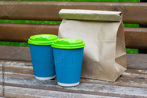 Paper bag with takeaway food and glasses with coffee on a bench in the park. Concept of food delivery, communication during quarantine covid 19 © queen1987