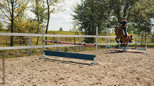 Female jockey on a dark bay horse jumping the obstacles in the sandy parkour. Horse Equestrian sports show jumping competition. Trees against a cloudy sky in the background.