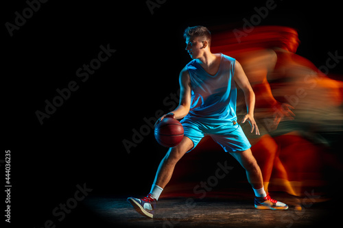 Young caucasian male basketball player playing basketball isolated over dark studio background in mixed light. Concept of healthy lifestyle, professional sport, hobby.