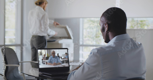 Back view of african employee sitting at desk in office having video chat with boss on laptop