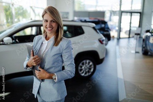 Female salesperson at a car showroom, holding a laptop and smiling to the camera. photo