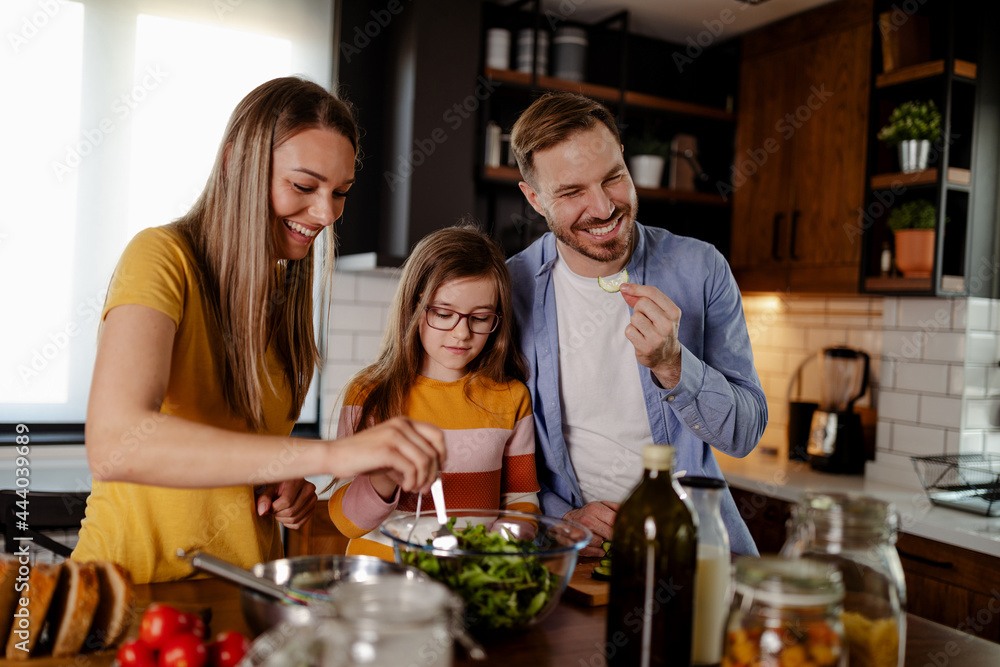 Cute little girl and her beautiful parents are smiling while cooking in kitchen at home.