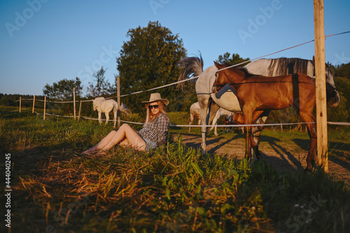 Beautiful young blonde woman dressed safari style in hat and plaid shirt posing with thoroughbreds horses on farm in sunset field outdoor at countryside. Horses behind an energized electric fence © Алина Троева
