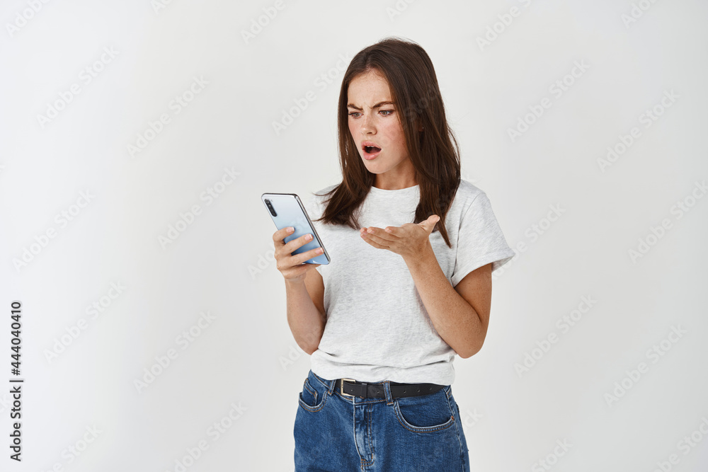 Young woman looking angry and confused at smartphone screen, complaining on broken app, standing over white background Young woman looking angry and confused at smartphone screen, complaining on broken app, standing over white background