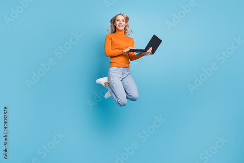 Photo of confident pretty young lady wear orange sweater jumping high chatting modern gadget smiling isolated blue color background