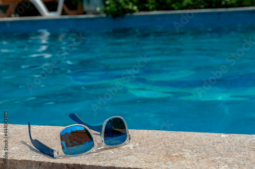 Blue fashionable sunglasses with mirror glasses lying on the stone floor with reflection of slide in them, pool water in background