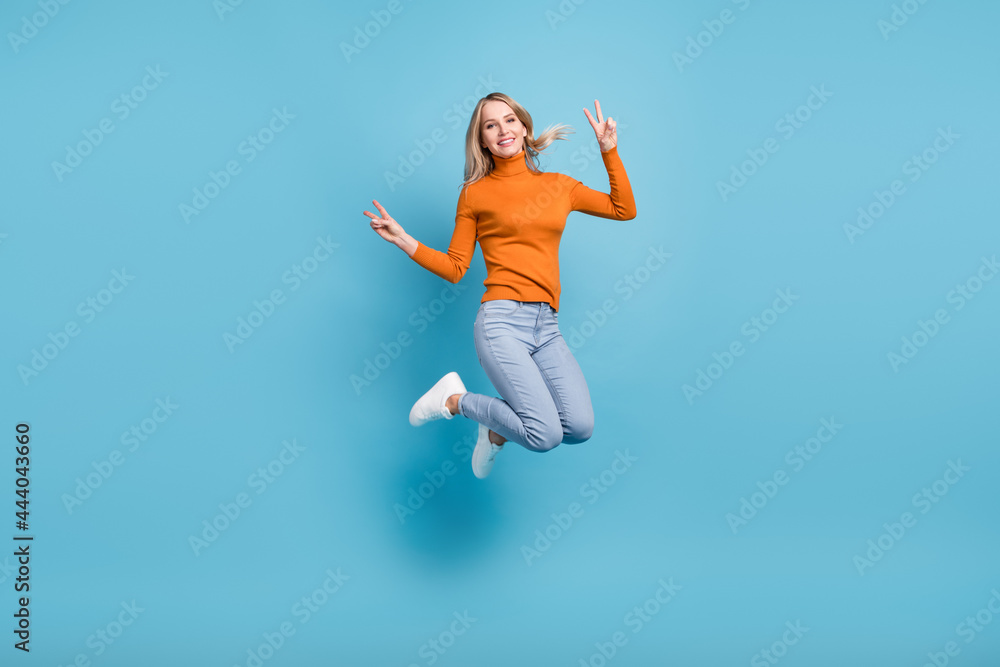 Photo of shiny sweet young woman dressed orange pullover smiling jumping high showing v-signs isolated blue color background Photo of shiny sweet young woman dressed orange pullover smiling jumping high showing v-signs isolated blue color background