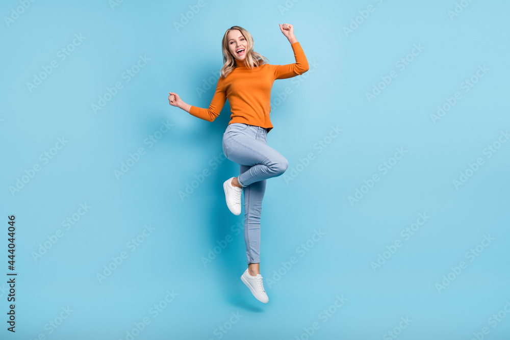 Fototapeta premium Photo of adorable lucky young lady wear orange sweater jumping high rising fists smiling isolated blue color background