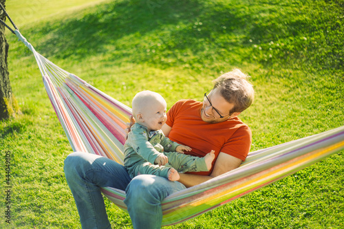 portrait of attractive nerd  man  with glasses in the park  with green lawn have a nice sunset with  a baby boy next to the hammock  . Happy fatherhood