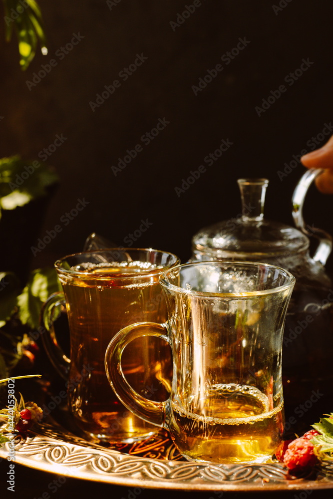 tea is poured into glass mugs. A teapot and a cup of tea stand on a copper tray next to raspberry leaves and berries