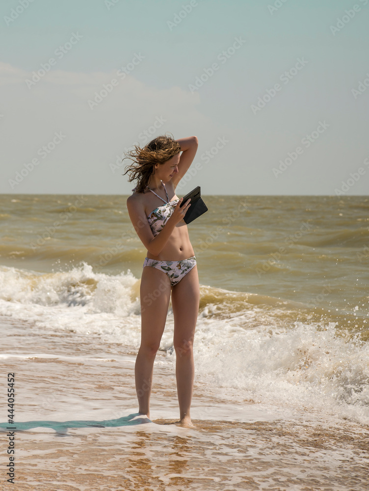 Naklejka premium a young slender girl stands on the seashore and poses for a photo with her phone