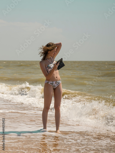 a young slender girl stands on the seashore and poses for a photo with her phone
