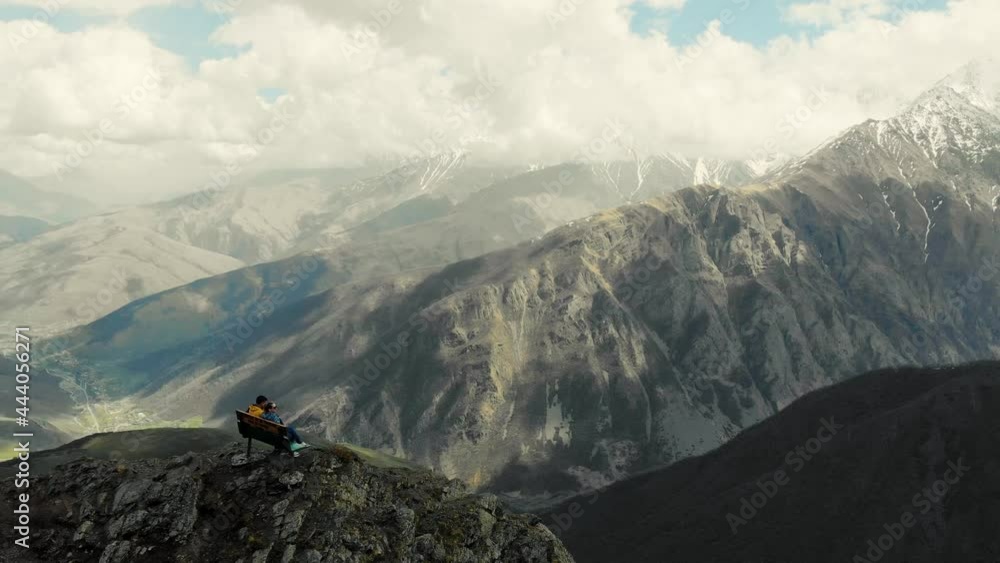 Couple sits on bench at top of mountain, the Caucasus. Bench stands on edge of mountain overlooking huge Caucasus mountains. Snowy peaks of the mountains. Clouds hang from above. Terrible beauty Couple sits on bench at top of mountain, the Caucasus. Bench stands on edge of mountain overlooking huge Caucasus mountains. Snowy peaks of the mountains. Clouds hang from above. Terrible beauty