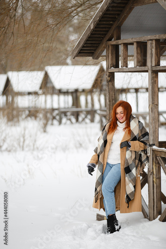 Happy woman in winter snow park wearing beige coat and large plaid scarf. Romantic walk in rustic style. Female in classic clothes with red hairs. Redhead smiling young girl near wood alcove or gazebo