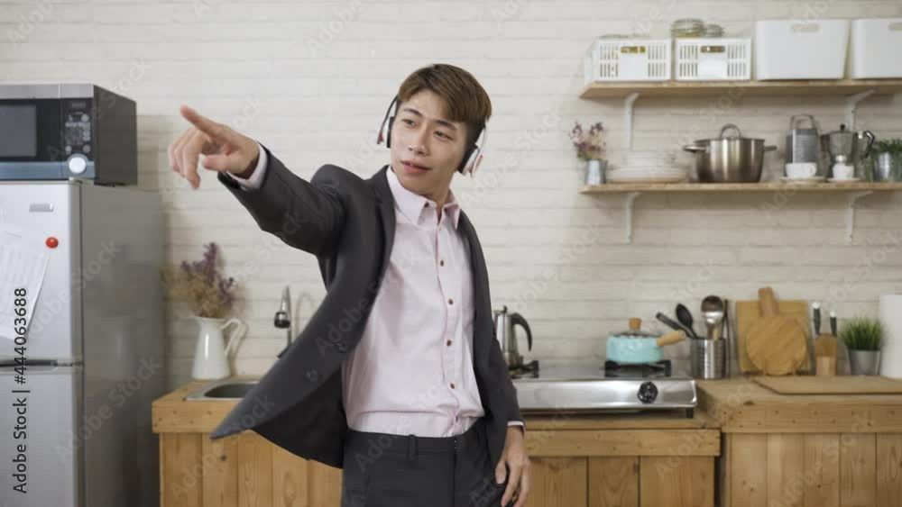 slow motion hyped asian businessman wearing earphones is pointing with index finger while having fun dancing to the music in the kitchen at home. slow motion hyped asian businessman wearing earphones is pointing with index finger while having fun dancing to the music in the kitchen at home.