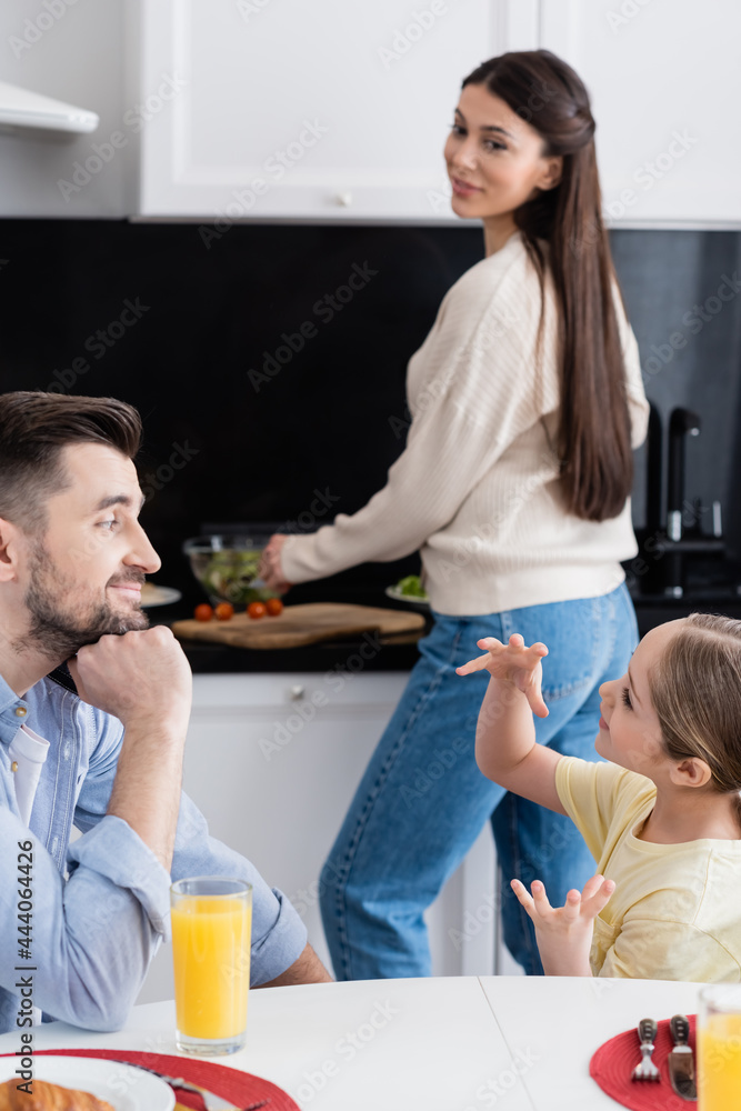 child gesturing while talking to smiling dad and blurred mother preparing breakfast in kitchen child gesturing while talking to smiling dad and blurred mother preparing breakfast in kitchen