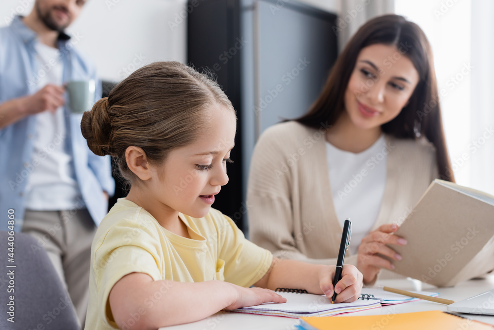 girl writing in notebook near blurred mom and dad with cup of coffee on background girl writing in notebook near blurred mom and dad with cup of coffee on background