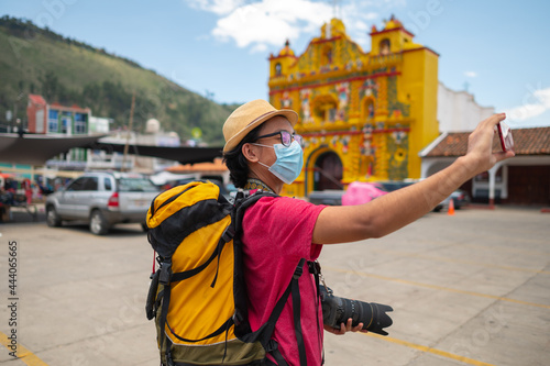 Portrait of a tourist with a medical mask, backpack, and camera taking a selfie with a cell phone.